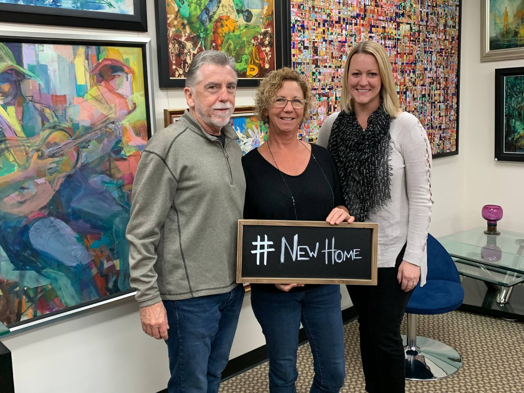 Jennifer Rigsby Standing at Closing with two Clients Jennifer Rigsby Standing with Clients at Closing Holding a Sign That Says #NewHome