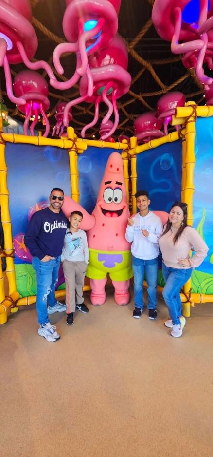 Maria Colon and her family pose with Patrick Star from SpongeBob SquarePants, smiling and enjoying a fun day together surrounded by colorful jellyfish decorations in Orlando, Florida.