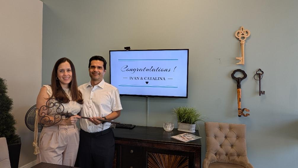 Maria Colon’s happy clients, Ivan and Catalina, celebrate their home closing at the Wemert Group Realty office in Orlando, Florida. They’re smiling while holding a large decorative key, with a “Congratulations! Ivan & Catalina” message displayed on the screen behind them.