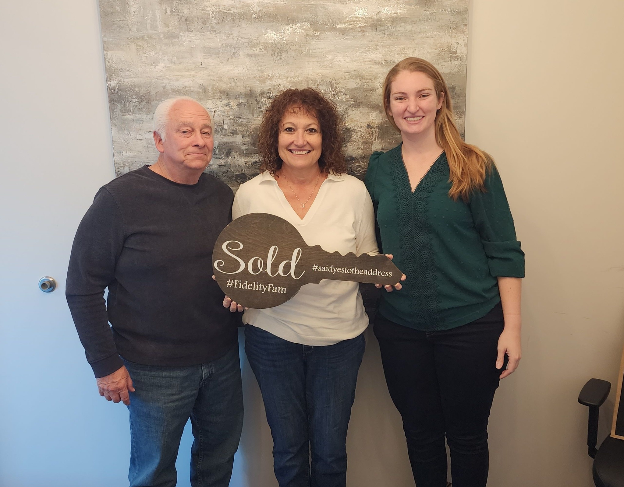 Orlando Realtor® Jessica Wicker celebrates with her clients on closing day as they hold a wooden “Sold” key sign at Fidelity National Title.