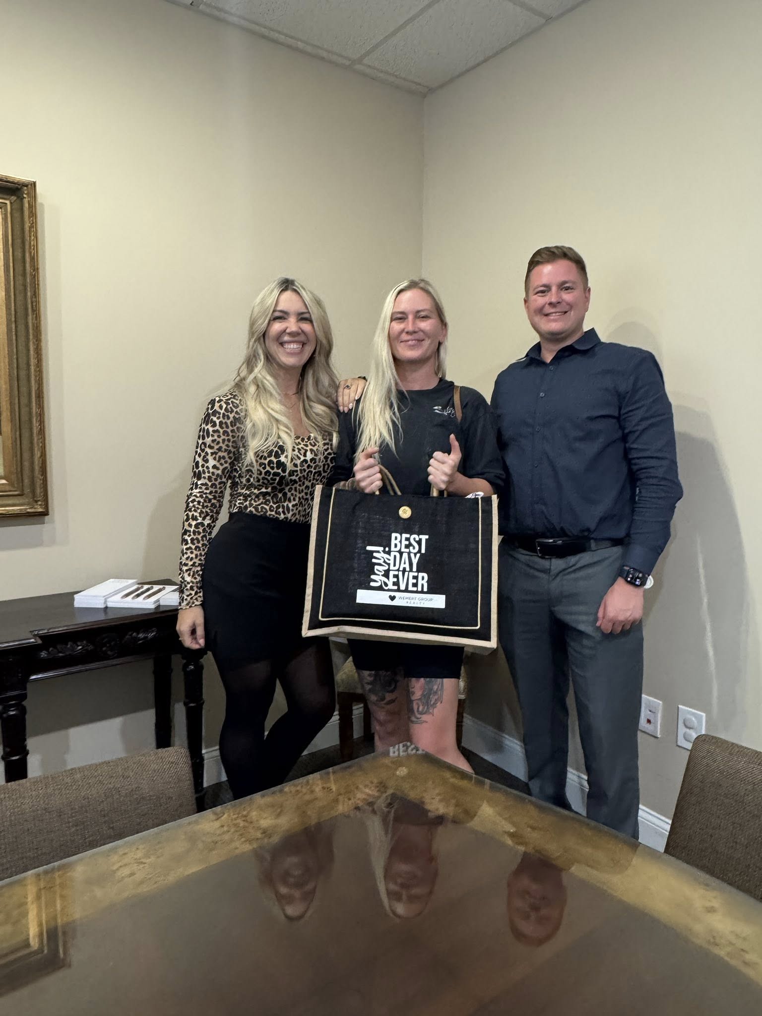 Team Tadasha Celebrates Closing Day with Client at Wemert Group Realty Two real estate agents from Team Tadasha stand with a smiling client on closing day at Wemert Group Realty. The client holds a black “Best Day Ever” closing gift bag while all three pose together in an office setting.